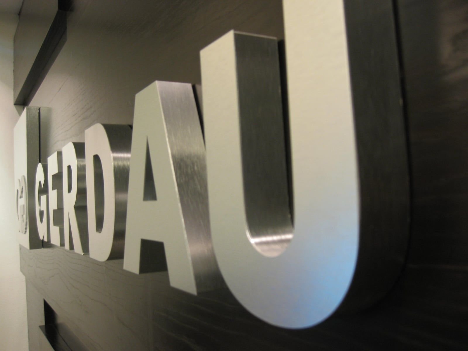 Letras 3D de aluminio natural cepillado con el logotipo de Gerdau sobre panel de madera oscura